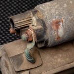 A close-up of a corroded solenoid valve. The valve is part of a corroded vehicle part, which sits on a table.