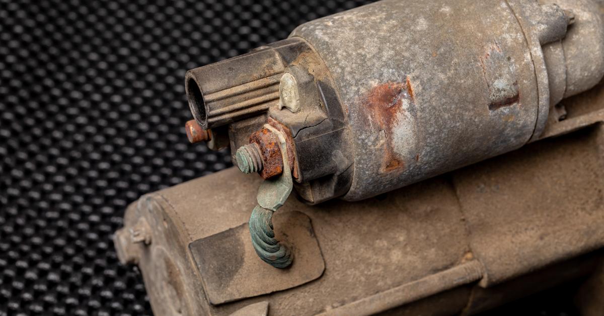 A close-up of a corroded solenoid valve. The valve is part of a corroded vehicle part, which sits on a table.