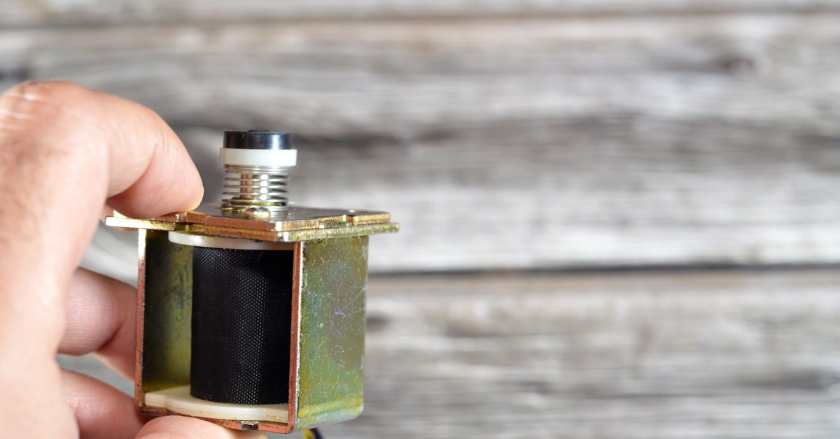 A close-up of a hand holding a solenoid valve. The valve is black and gold with a few wear and tear marks.