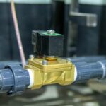 A close-up of a black and gold solenoid valve attached to gray pipes in a factory. The background is blurred.