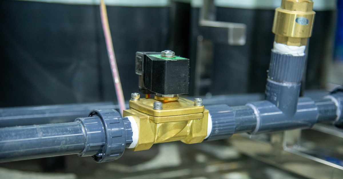 A close-up of a black and gold solenoid valve attached to gray pipes in a factory. The background is blurred.