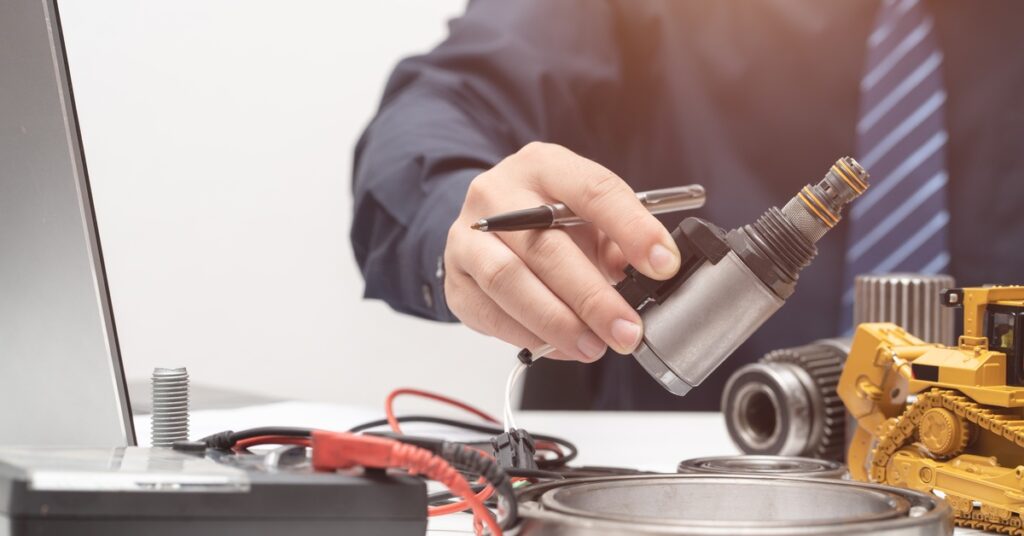 A close-up of a man sitting at a desk near a laptop and engineering components. He holds a solenoid valve and a pen.