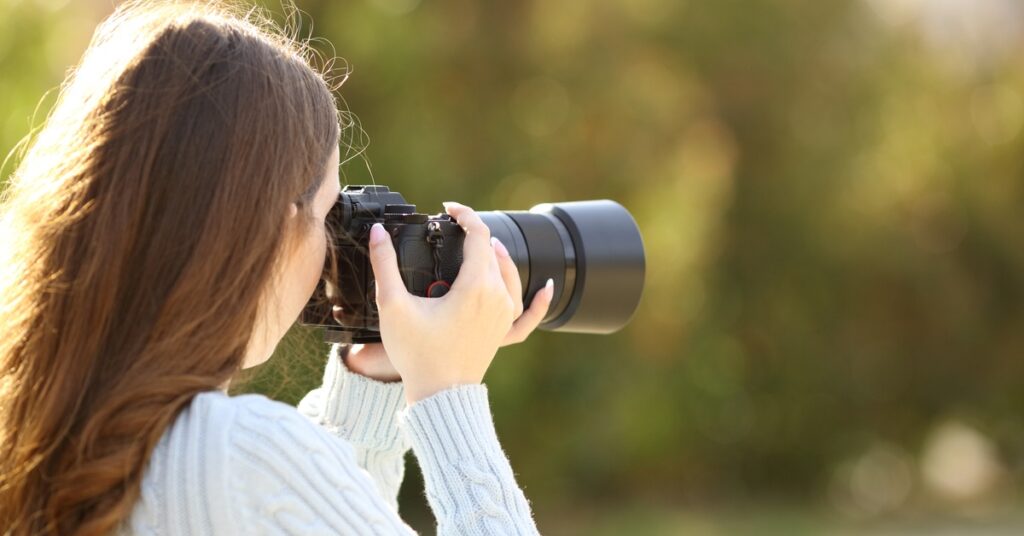 A side view of a woman taking pictures outside with a camera. She has brown hair and a light-colored sweater.