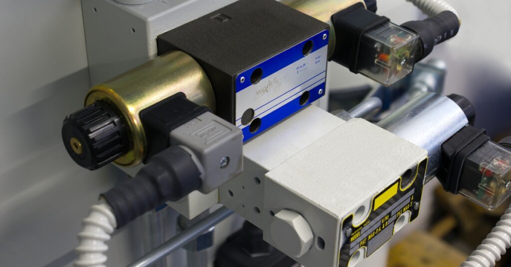 An industrial solenoid valve with a black and blue label connects to multiple cables and pipes on a mounted panel.