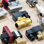 Various brass and metal solenoid valves sit on a light-colored surface. They are arranged in rows.