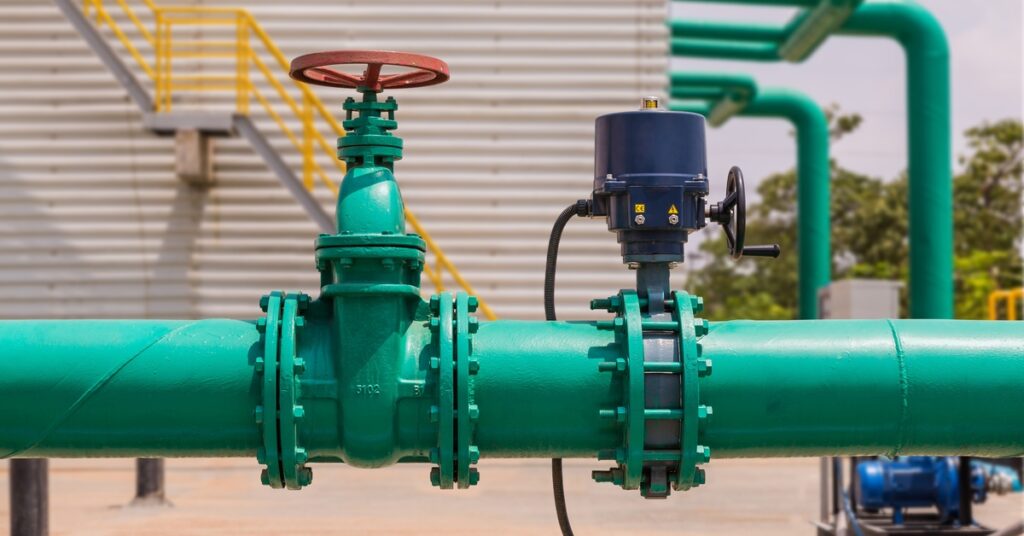 A green pipeline with a valve and actuator sits near a building with metal siding and yellow stairs.