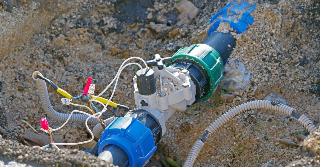 Wiring and couplings surround an exposed underground pipe with a solenoid valve in sandy soil at an active installation site.