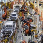 Robotic arms are assembling silver car bodies inside a large vehicle production line with orange railings.