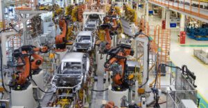 Robotic arms are assembling silver car bodies inside a large vehicle production line with orange railings.