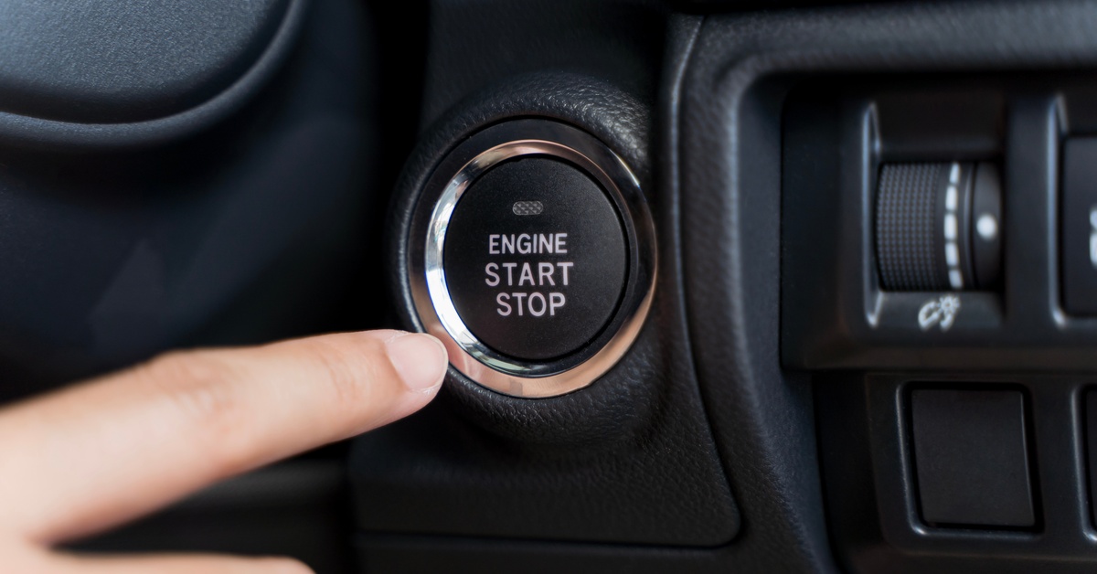 A finger is pressing a circular engine start-stop button beside climate controls inside a black car dashboard panel.