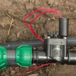 An irrigation solenoid valve with red wiring connecting black plastic pipes using green fittings resting on soil.