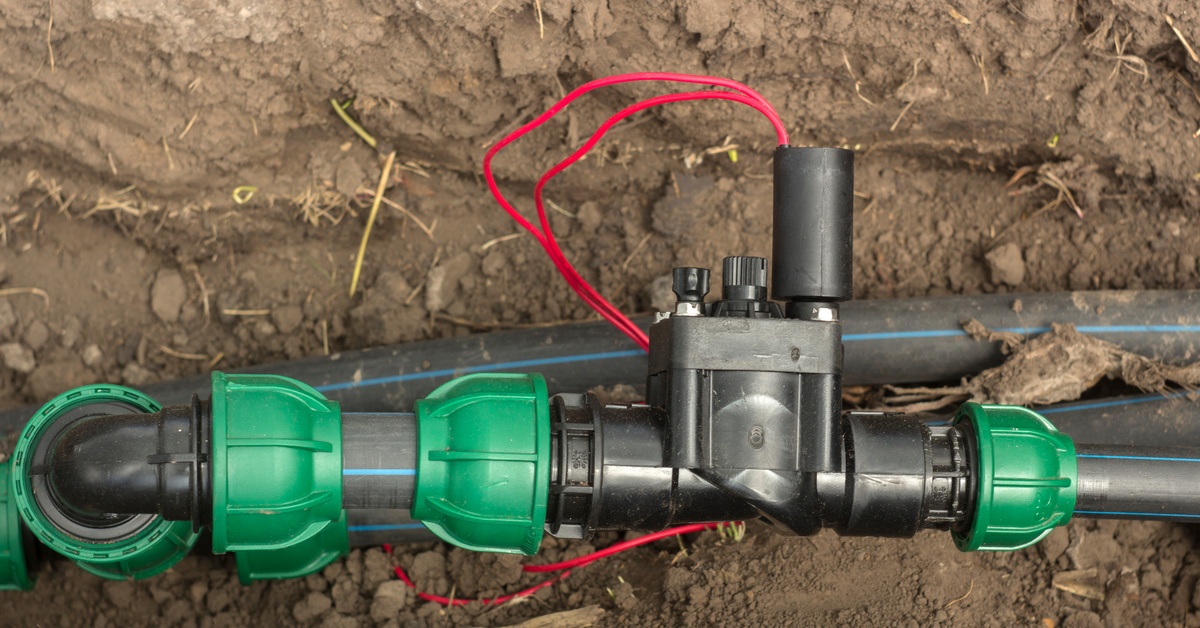 An irrigation solenoid valve with red wiring connecting black plastic pipes using green fittings resting on soil.