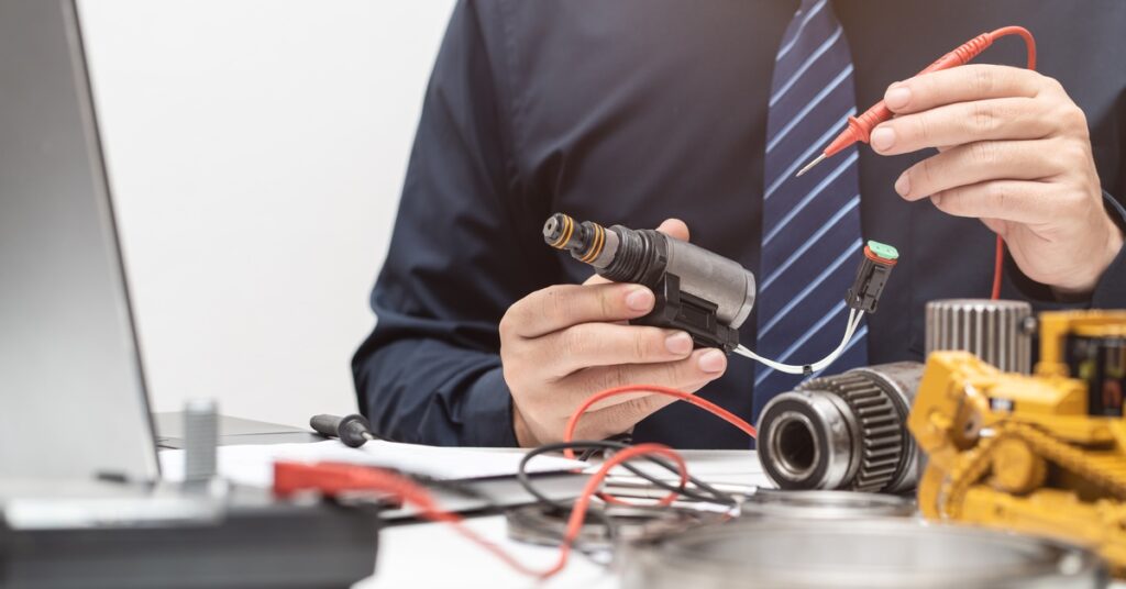 A man in a dark shirt and striped tie holding probes while testing engine wiring near a cluttered desk.