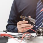 A man in a dark shirt and striped tie holding probes while testing engine wiring near a cluttered desk.