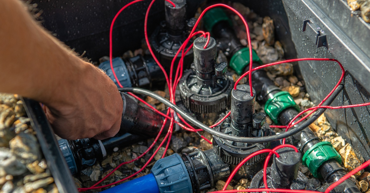 A person reaching into an open irrigation valve box with red wires and plastic pipe connectors on gravel.