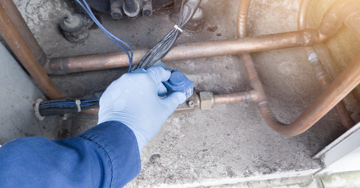 A gloved hand adjusting a blue valve connected to copper pipes, wires, and tubing inside a dusty system panel.