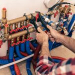 A person in a plaid shirt using a wrench to adjust multiple red and blue water pipes on a manifold panel.