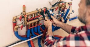 A person in a plaid shirt using a wrench to adjust multiple red and blue water pipes on a manifold panel.