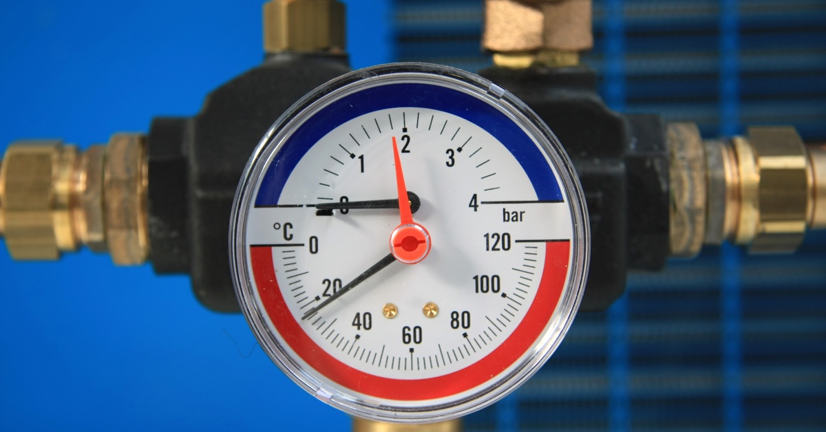 A round pressure and temperature gauge with red and blue markings mounted between brass pipes on a blue background.