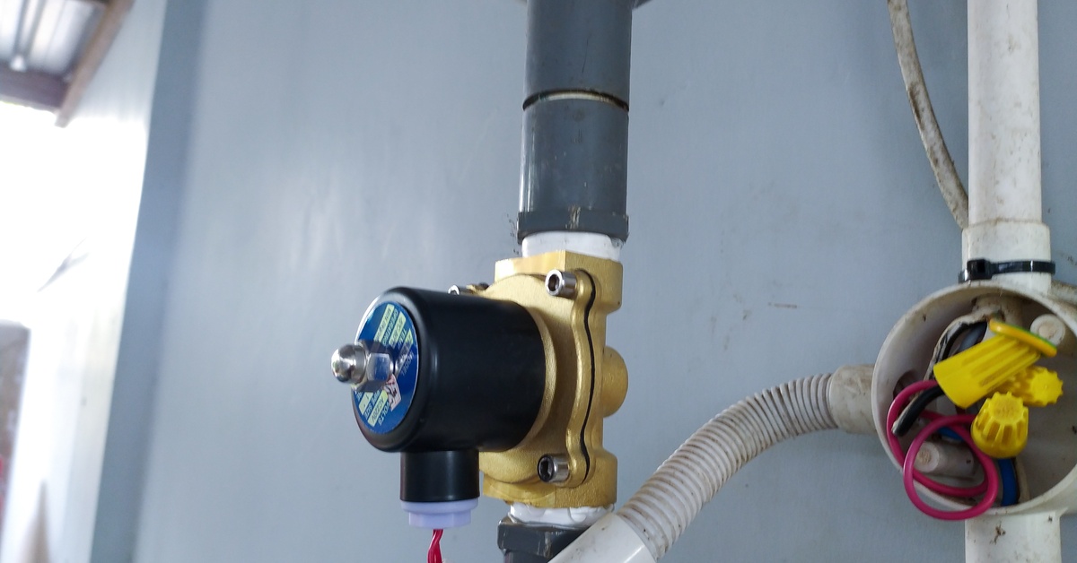 A black and brass solenoid valve connected to gray PVC piping and exposed wires beside a junction box on a wall.