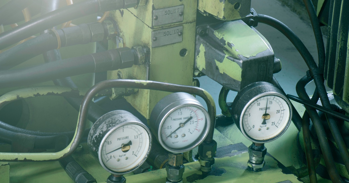 Three analog pressure gauges mounted on a worn green hydraulic control unit with connected hoses in an industrial setting.
