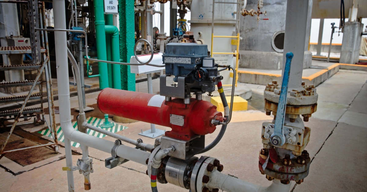 A red pneumatic actuator and solenoid on a white pipeline in an outdoor processing plant with various pipes and equipment.