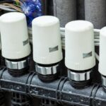 A row of white valve actuators mounted on a black manifold with insulated pipes and connected electrical wiring above.