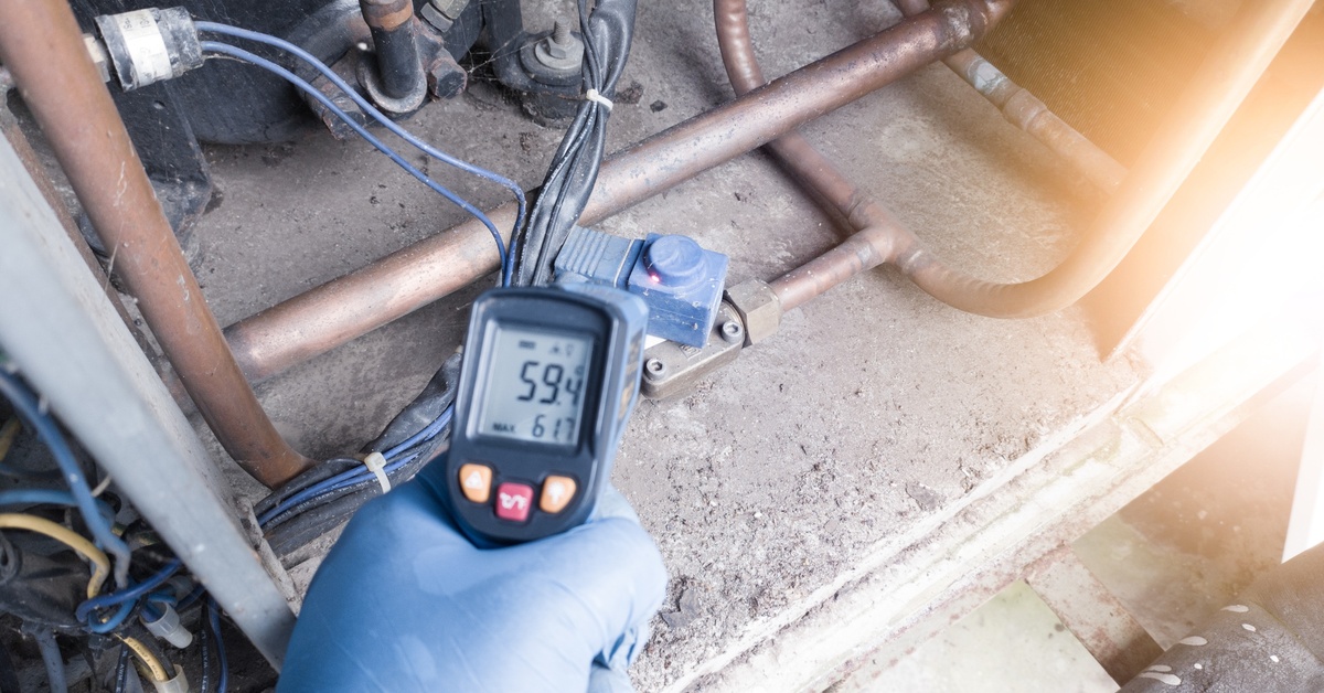 A person holding a digital infrared thermometer near copper pipes and wiring connected to HVAC equipment.