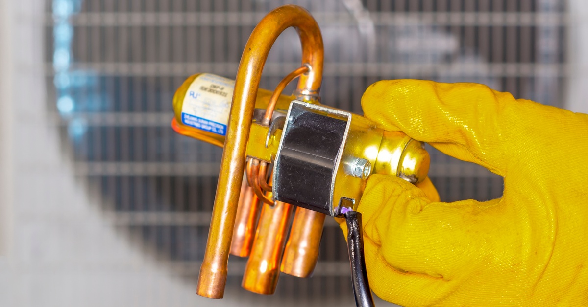 A person wearing gloves holds a copper solenoid valve component with wires in front of an air conditioning unit.