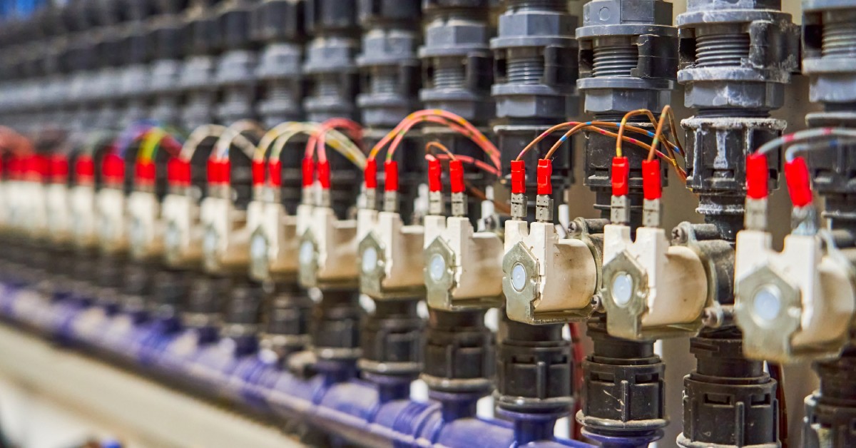 A close-up view shows several brass solenoid valves lined up with plastic gray and black parts attached to the top.