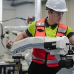 Two team members wearing yellow shirts, orange safety vests, and white hard hats work on white robotic arms.