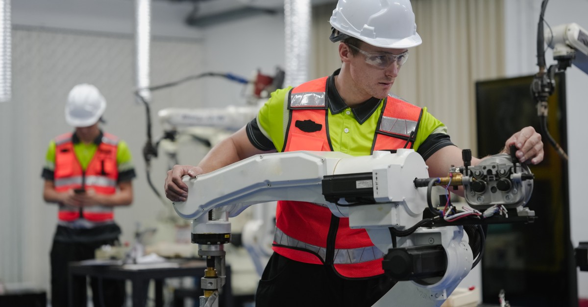 Two team members wearing yellow shirts, orange safety vests, and white hard hats work on white robotic arms.