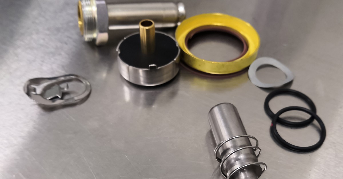 A dismantled solenoid valve sits on a stainless steel counter. The valve contains yellow, black, and silver pieces.