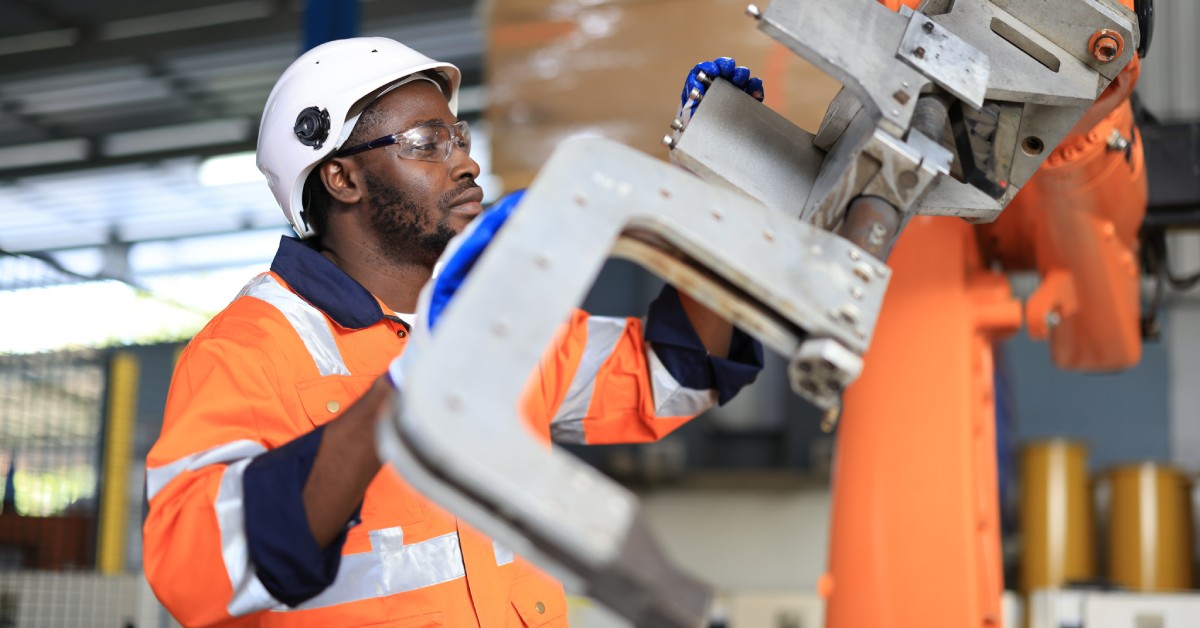 A man wearing an orange high-visibility jumpsuit works on an assembly line, holding a piece of metal.