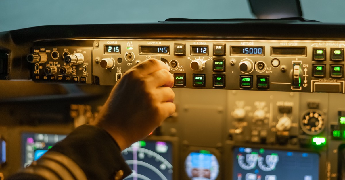 A close-up view shows a hand turning one of the many knobs on a flight control system. The system is illuminated.
