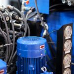 A close-up of an industrial hydraulic system with a blue motor, pipes, hoses, and pressure gauges mounted on machinery equipment.