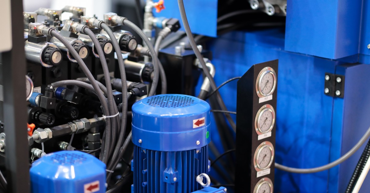 A close-up of an industrial hydraulic system with a blue motor, pipes, hoses, and pressure gauges mounted on machinery equipment.
