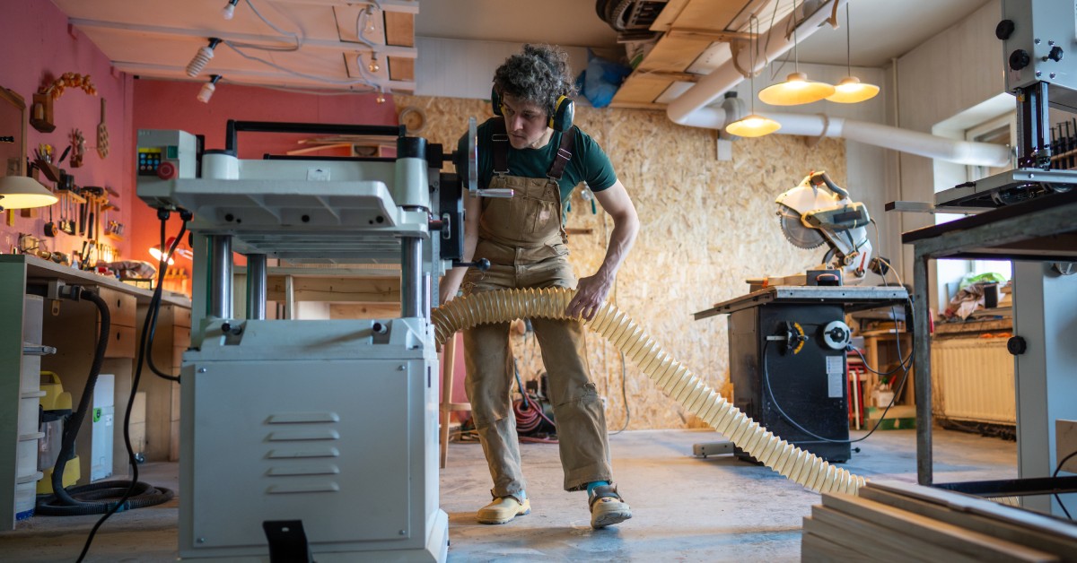 A person in a workshop using a large woodworking machine. They are wearing protective earmuffs and overalls for safety.
