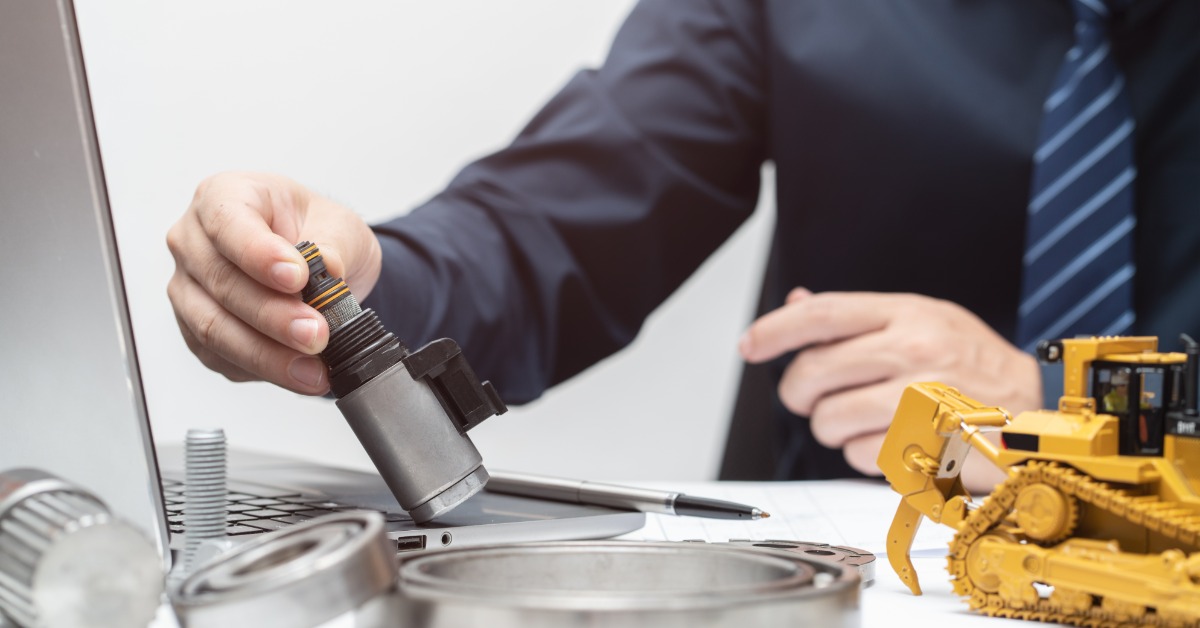 A person wearing a black button-down shirt and a blue tie is holding a black solenoid valve beside a toy bulldozer.