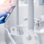 A person wearing a white lab coat and blue latex gloves holds a tablet while standing in a room with metal cylinders.
