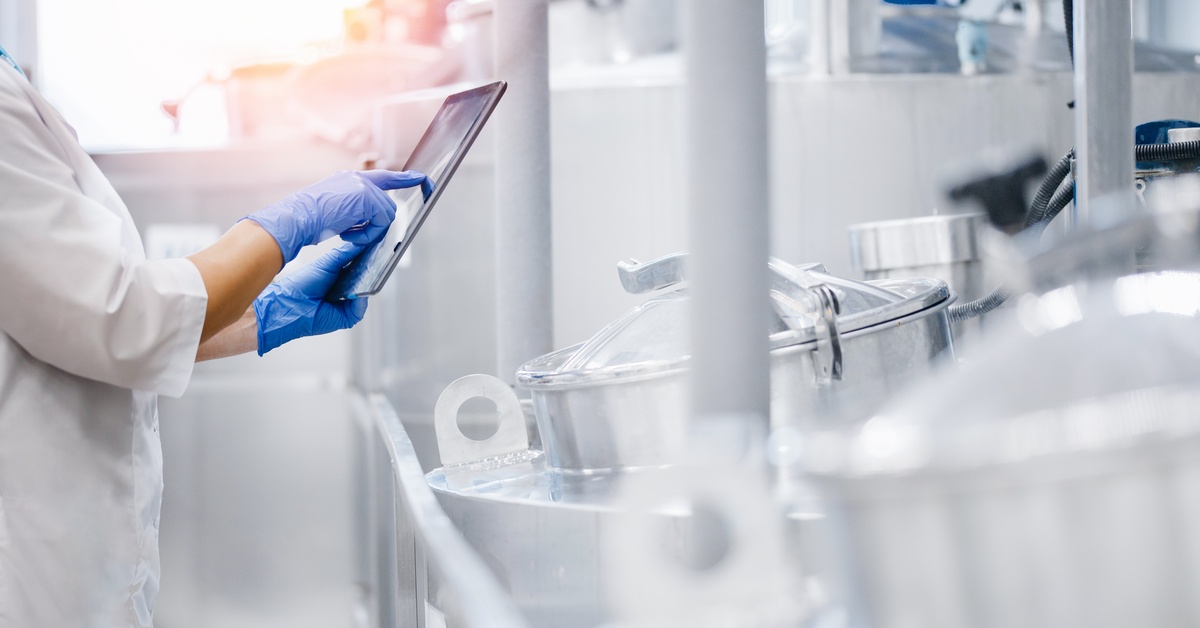 A person wearing a white lab coat and blue latex gloves holds a tablet while standing in a room with metal cylinders.