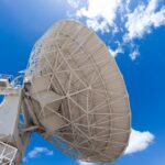 An oversized, white satellite dish is illuminated by the sun. The blue sky with a few white clouds is behind it.