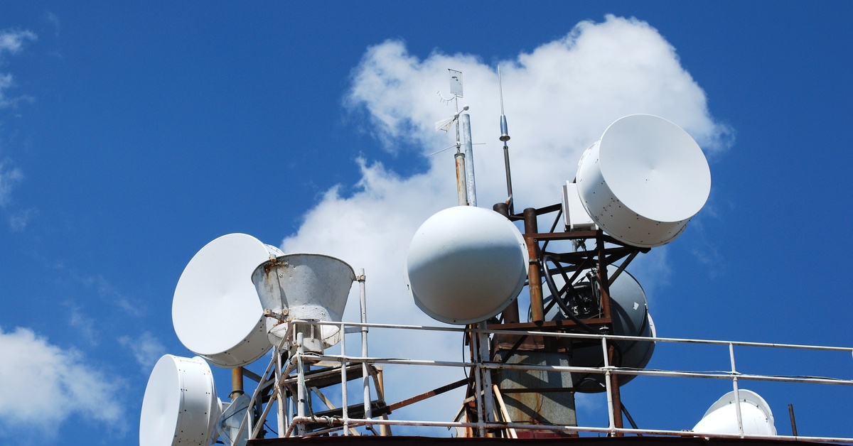 Several styles of satellites sit on top of a red building. The blue sky appears behind them with a few white clouds.