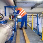 A man wearing a blue jumpsuit, a blue hard hat, and an orange safety vest is checking on industrial valves and pipes.