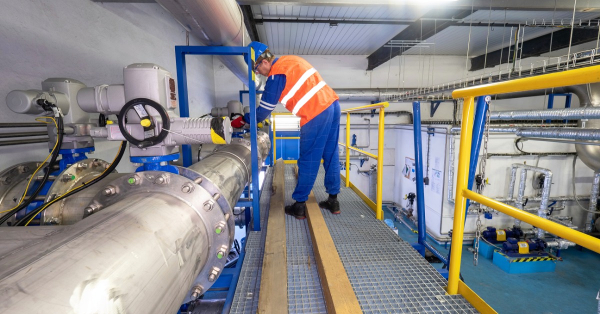 A man wearing a blue jumpsuit, a blue hard hat, and an orange safety vest is checking on industrial valves and pipes.