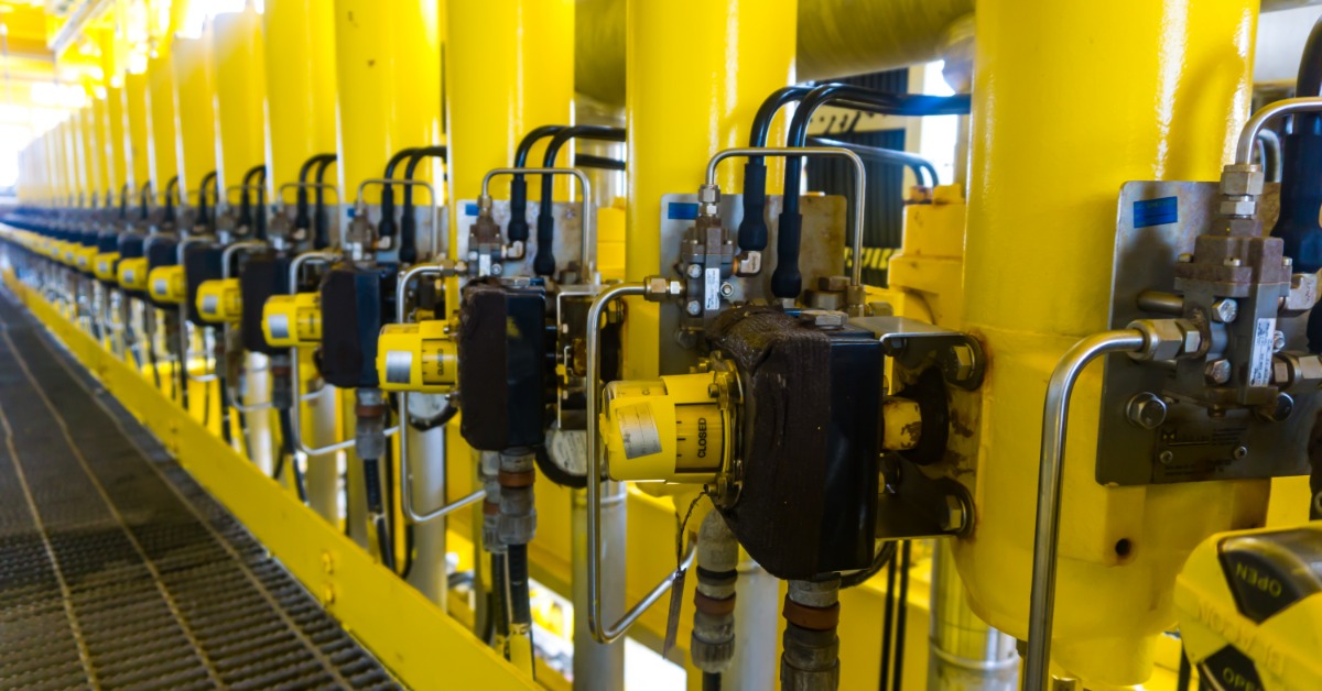 A long-line of bright, yellow pipes features a black solenoid valve on the front. The pipes are in an industrial setting.
