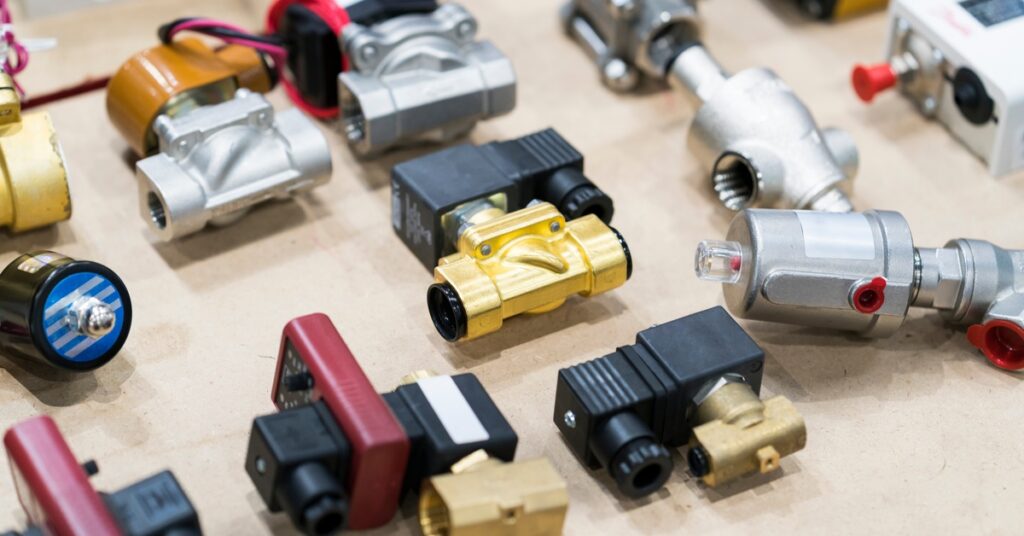 Various types of solenoid valves are scattered on a brown surface. The valves are bronze and attached to black plastic.