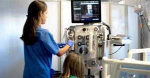 Two female medical professionals are working on a dialysis machine. One woman is standing, and one is squatting.