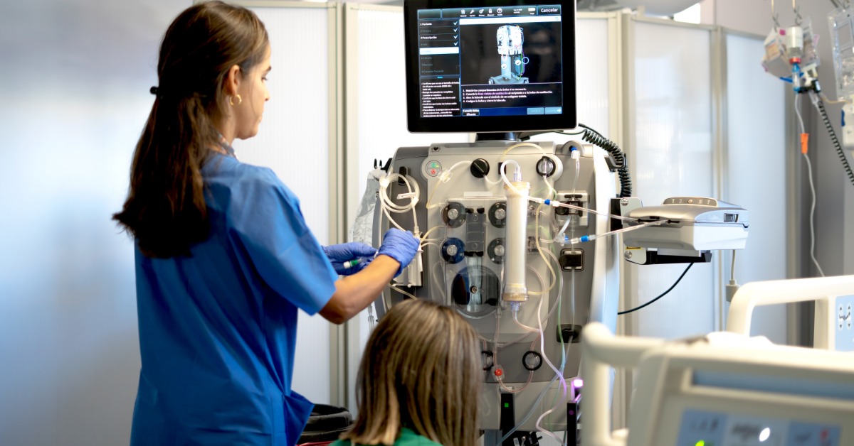 Two female medical professionals are working on a dialysis machine. One woman is standing, and one is squatting.