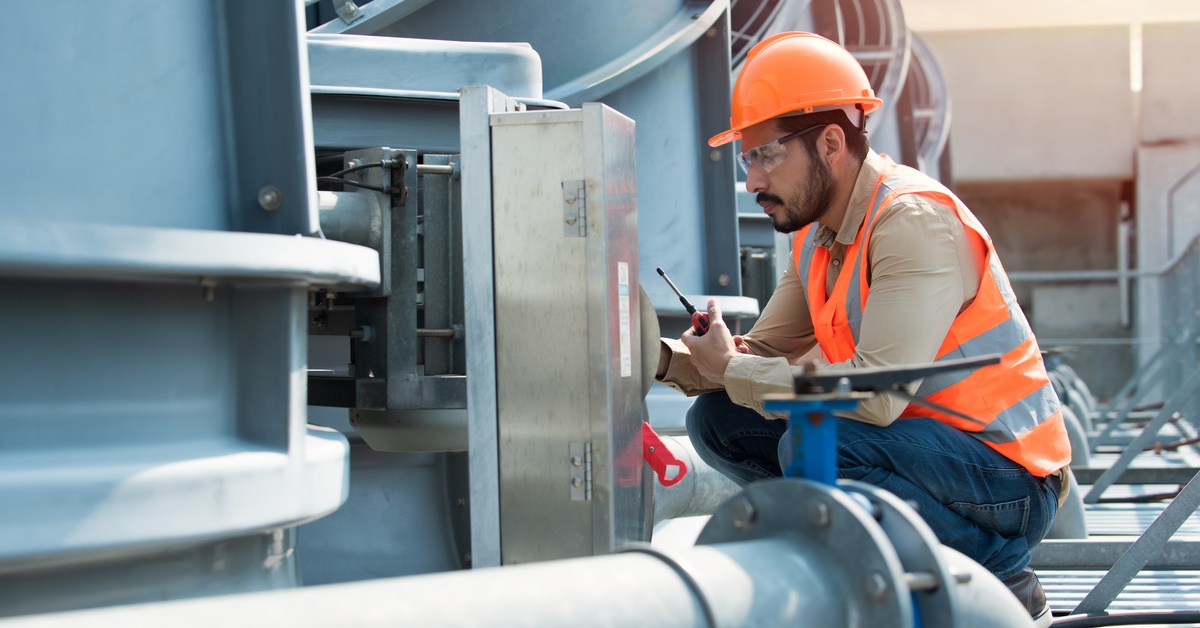 A man wearing a bright orange safety vest and helmet is checking on the control panel of an HVAC system.